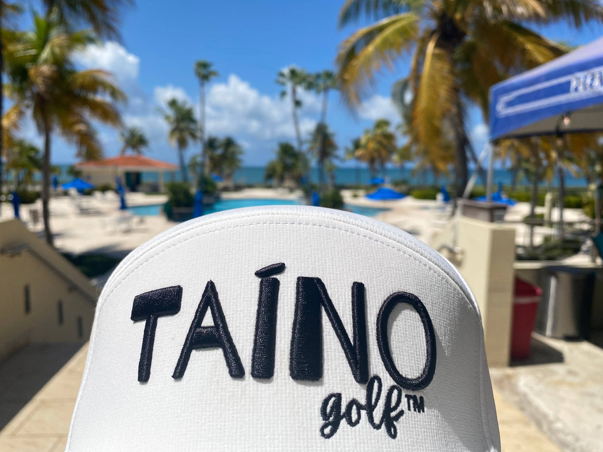 Taino Golf hat pictured in front of the pool at Costa Caribe Resort and Golf Course with the ocean and palm trees in the background.