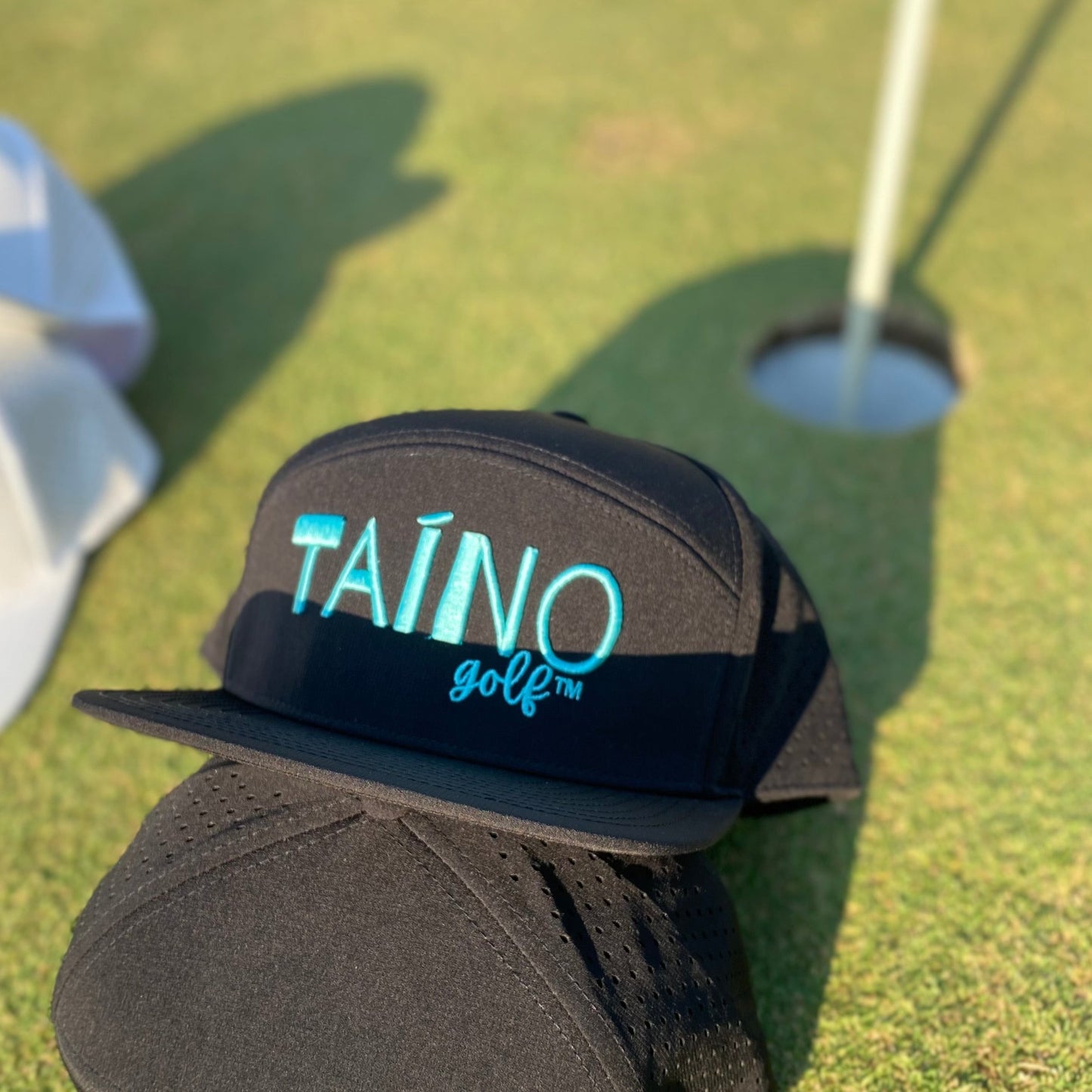 Waterproof Taino Golf hat in black with teal logo embroidered on the front panel, displayed on a putting green.