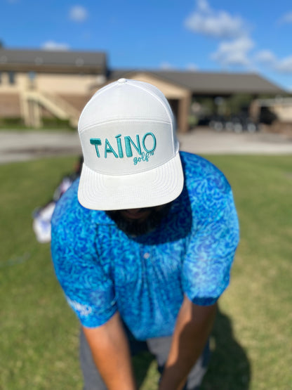 Golfer with a white Taino Golf hat with teal logo embroidered on the front, chipping at a golf course.