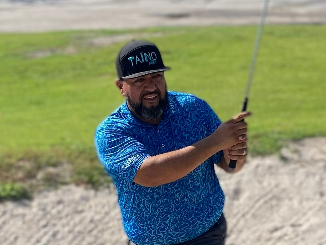 Golfer hitting out of bunker using a black Taino Golf hat with teal logo embroidered on the front of the hat.